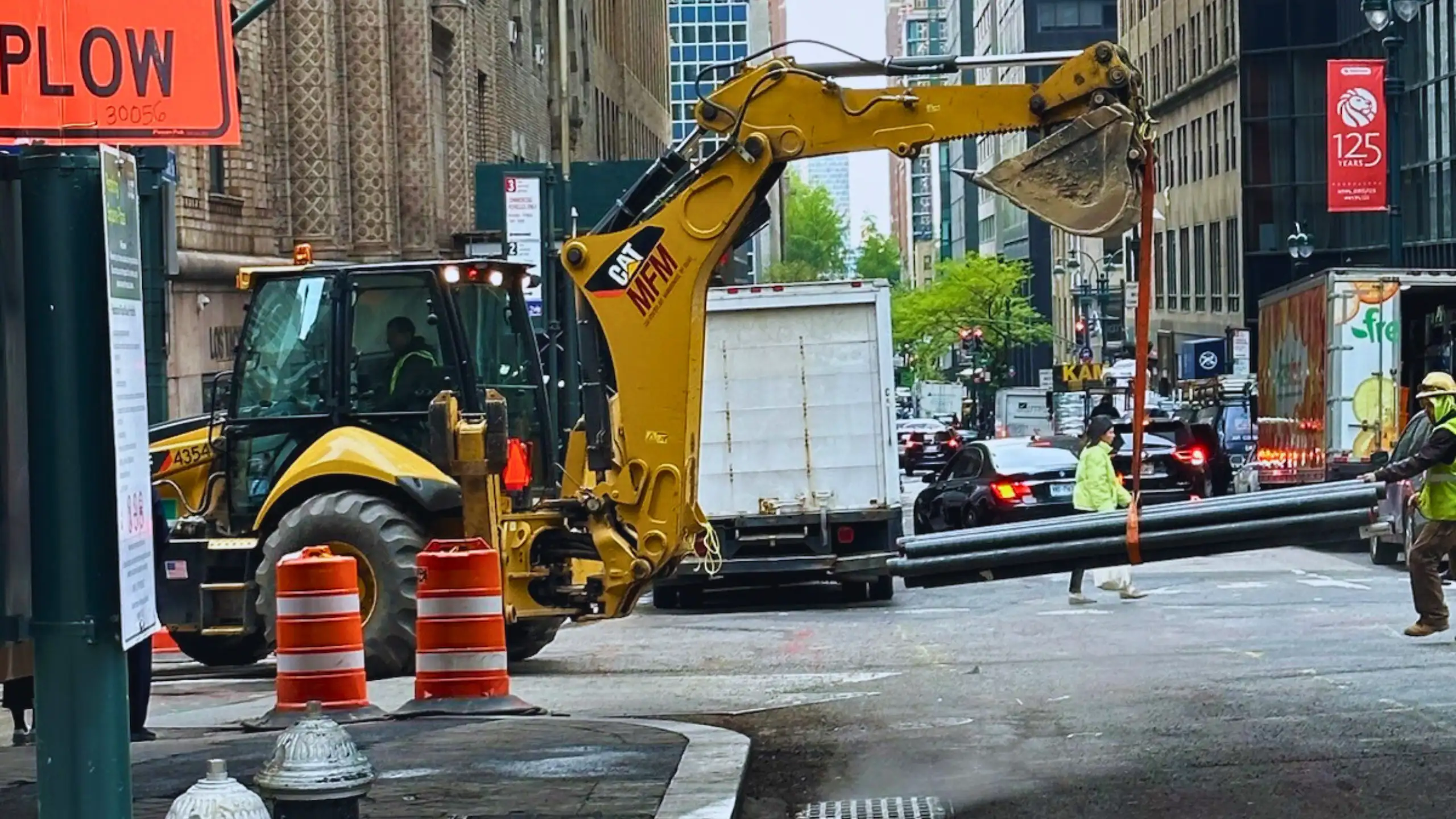 Yellow excavator lifting heavy pipes on a city street.