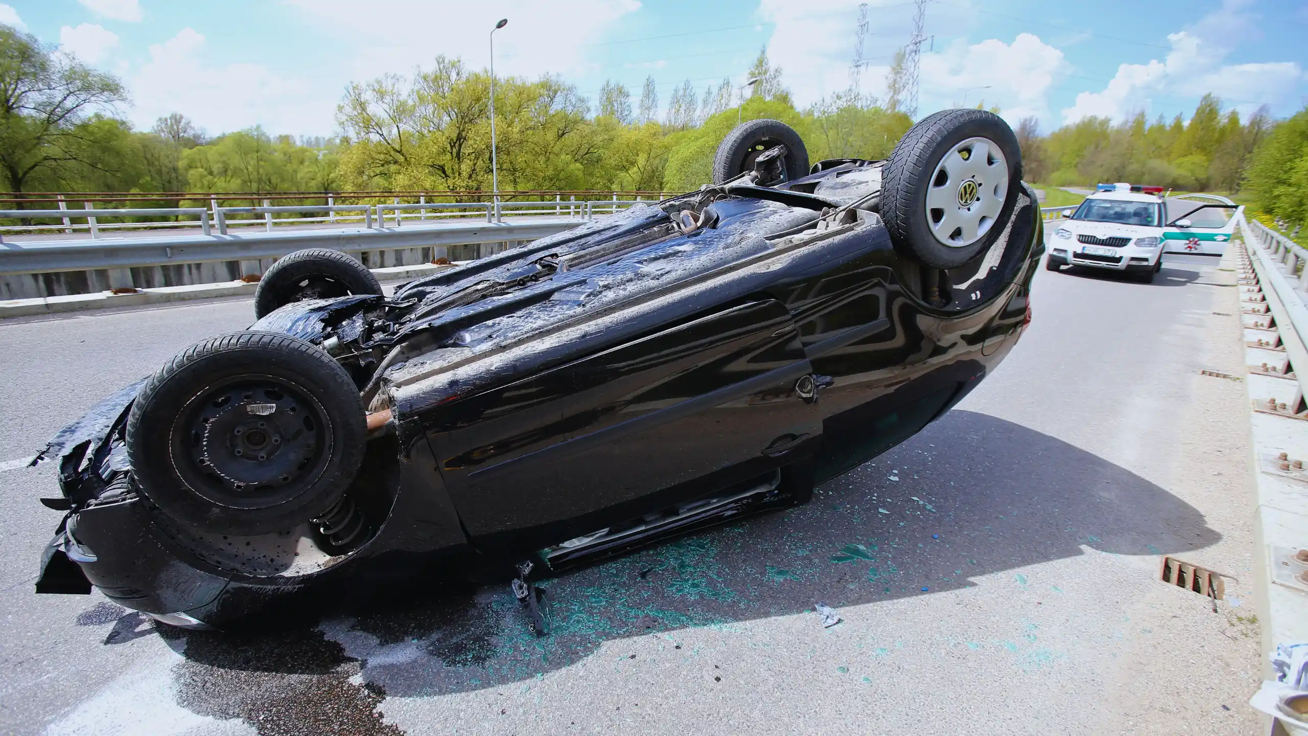 A black car is flipped upside down on a bridge after an accident, with a police car visible in the background.