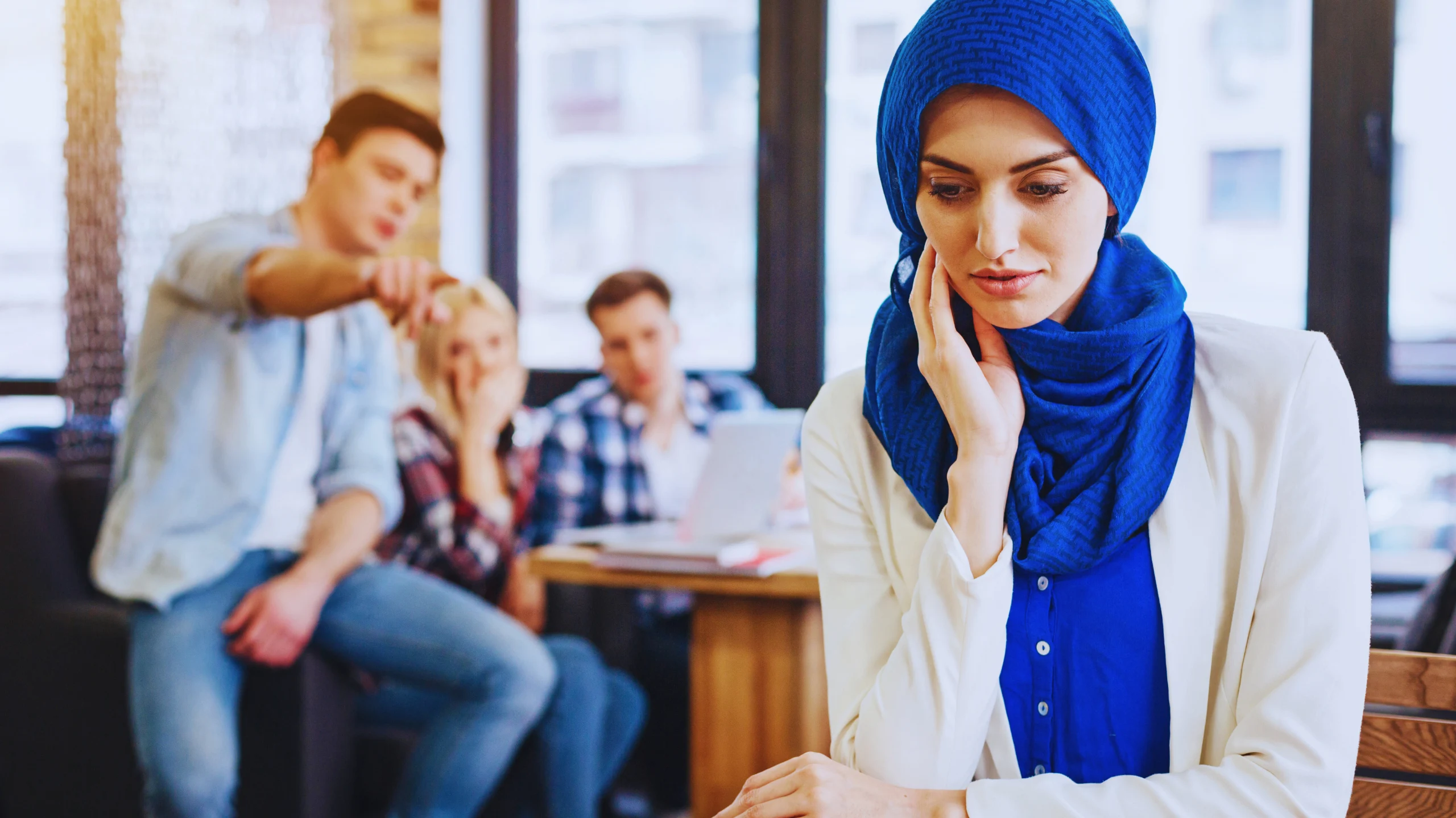 Woman wearing a blue hijab looks down thoughtfully while a group of people talk and gesture in the background.