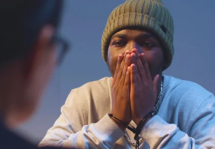 A handcuffed man in a beanie covering his mouth while being questioned by a blurry person.