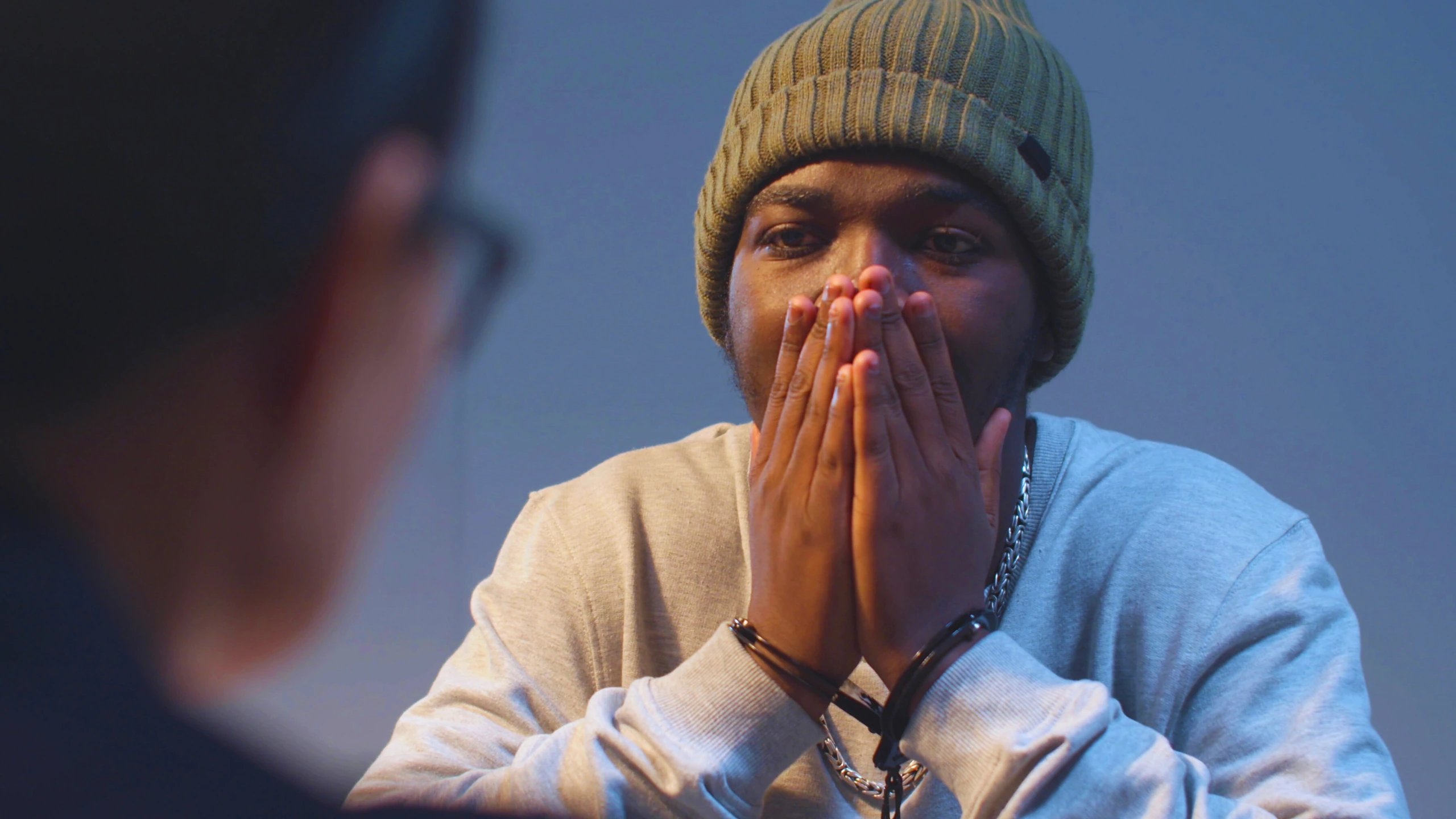 A handcuffed man in a beanie covering his mouth while being questioned by a blurry person.