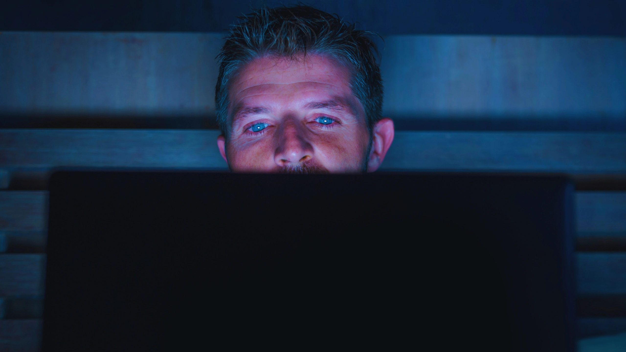 Man looking at a laptop screen in a dark room with blue light.